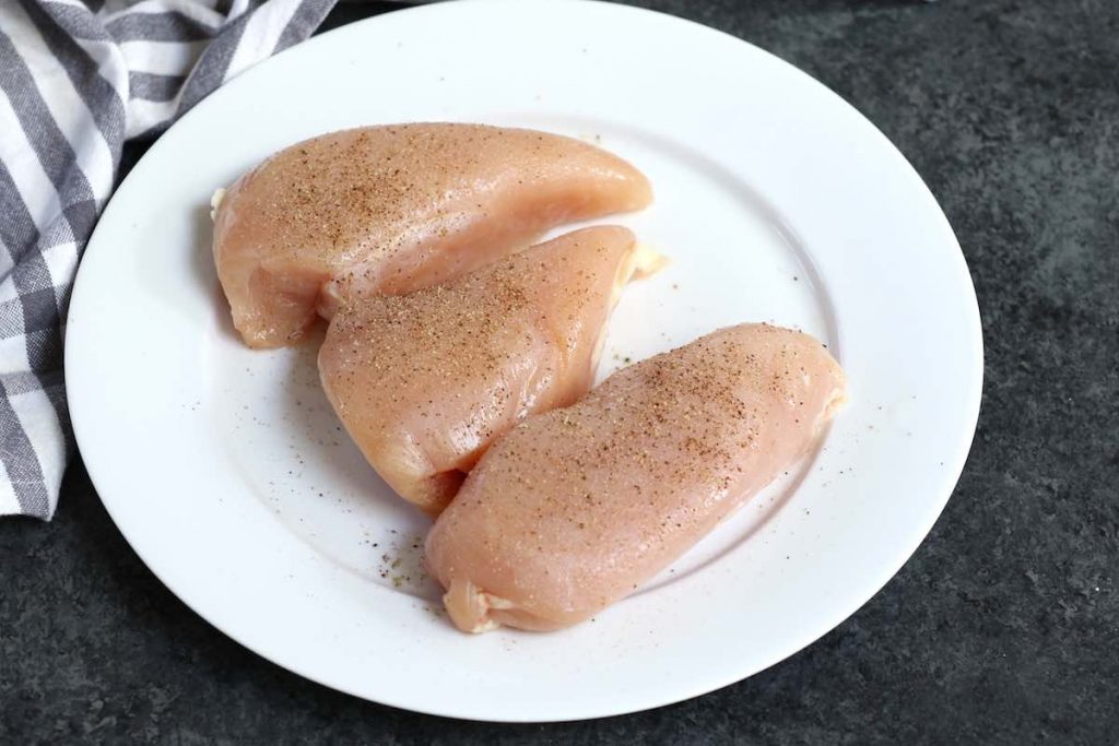Des blancs de poulet crus assaisonnés de sel, de poivre et de poudre d'ail sur une assiette blanche.